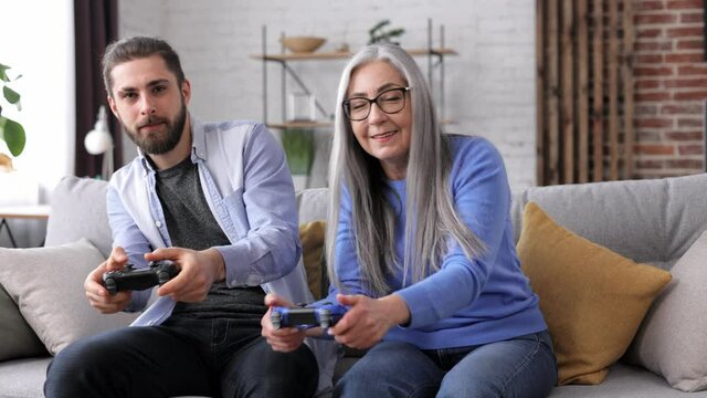Adult son and senior mother enjoy spending time together. Grandmother and grandson playing video games using gamepads at home. Mothers Day. Two generation family. Active modern elderly people.