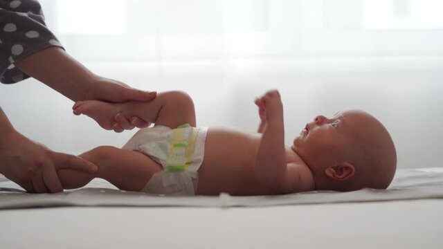Mother with baby boy doing exercise simple exercises to make baby bones and muscles stronger, newborn infant lying on bed against the window. High quality 4k footage