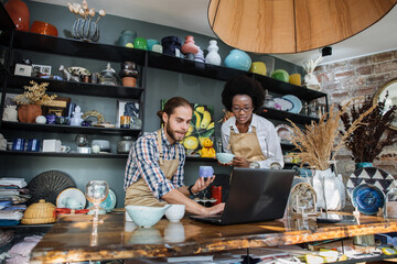 Two multicultural people working on modern laptop during inventory process at decor store. Young caucasian man and african woman rechecking all assortment at shop.