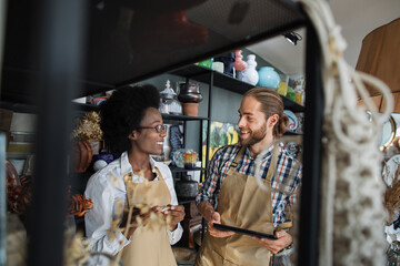 Smiling african woman and caucasian man standing together at decor store with digital tablet in hands. Two coworkers in beige apron doing inventory at shop.