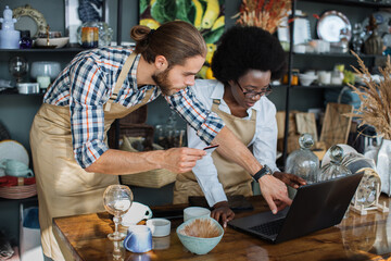 Two multicultural people in uniform analysing clients orders made on website of decor store. Handsome man holding credit card, charming woman typing on laptop.