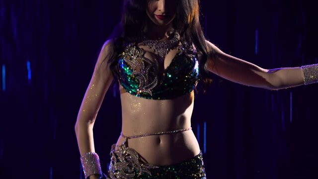 Young woman in an exotic green suit performs belly dance, moves hips and wriggle half naked body. Close up of dancer in traditional oriental costume amid falling drops of rain, smoke and blue light.