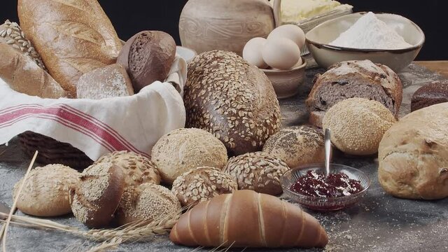 Different types of fresh baked Dutch bread is on the wooden table. wicker basket stands on the side.  There are also eggs in the clay bowl, flour and butter on the back, a vase with jam. Background is