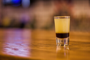 A tequila shot of cocktail composed of different syrup in layers in a small glass on wooden table in a bar. Art of alcohol liquer drink making. selective focus with blur background.