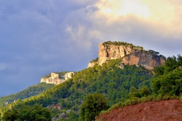 Fototapeta na wymiar Orange massive rock on green mountainside covered with trees, climbing sector. Blue sky with white clouds. Beautiful nature, mountains. Siurana, Catalonia, Spain.