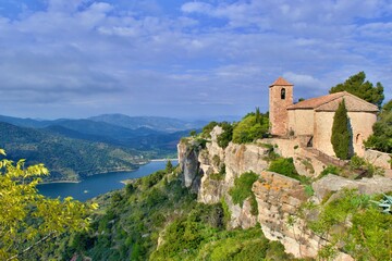 Fototapeta na wymiar Romanesque church of Santa Maria de Siurana on top of rock, view into valley, mountains on the background. Blue sky with white clouds. Siurana, Catalonia, Spain.