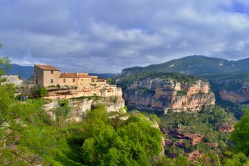 Fototapeta na wymiar Inn, building, house on the edge of cliff with beautiful view into the valley of red rocks, green bushes. Mountains in the background. Sun, clouds. Siurana, Catalonia, Spain.