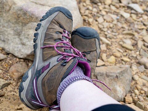Close up picture of a pair of brown and pink hiking boots with purple wool socks with a rocky sandy background, ready for adventure and trails.