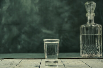 Vodka in decanter and shot glass on wooden plank table. Atmosphere bar drinking. Copy space for text on dark wall. Concept solitary alcoholism © Koirill