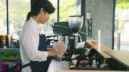 Young barista man in apron cleaning and preparing bar for visitors working in coffee shop. Cheerful young man waiter cleaning table and working in coffee shop.