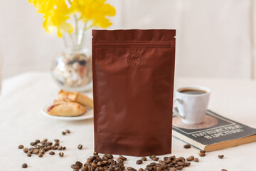 Blank coffee packaging, coffee seeds, cup with coffee on old book, yellow flowers and cookies, coffee packaging mockup with empty space to display your branding design.