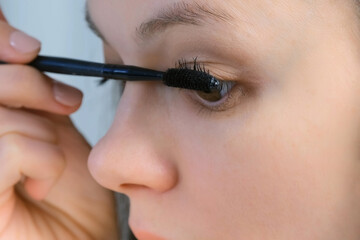 Woman is painting lashes using mascara and brush looking at small mirror, closeup face view. Female is doing makeup. Everyday routine for woman.