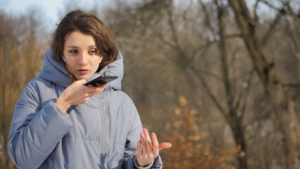 Lady in blue coat is sending audio voice message explaining something on smart phone at outdoor talking to mobile assistant. Girl using smartphone voice recognition and dictation