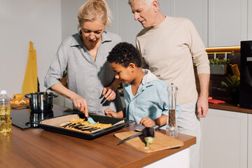 Caring grandparents teaching little boy cooking, while showing how greasing vegetables