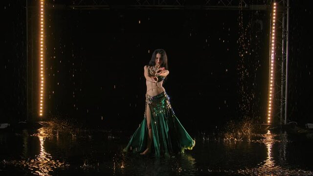 Attractive belly dancer shakes hips in shining oriental costume amid raindrops on surface of the water. Brunette dancing oriental dance on black studio background with bright lights. Slow motion.