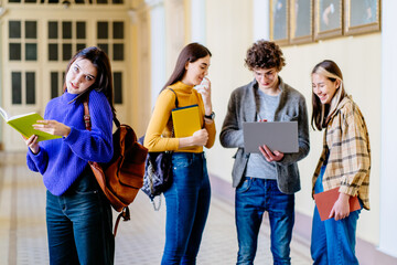 Positive young woman university student with backpack and books talking on cell phone, looking at camera, smiling with friends stand in a university hall during break on background.