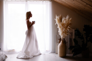 Early awakening morning magic. woman in sheet stands by the window drinking coffee from a mug. Slow...
