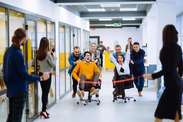 Friendly work team  ride chairs in office room cheerfully excited diverse employees laugh while enjoying fun work break activities, creative friendly workers play a game together.