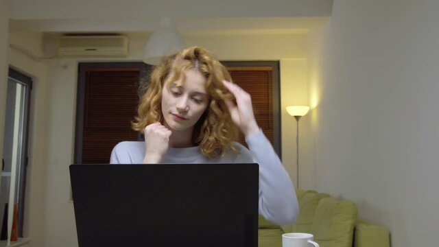 Cute girl sitting on a table in living room, infront of a laptop computer, drinking coffee. Portrait of a womam working from home. Camera moving left.