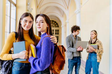 Two attractive girl friends wearing yellow and violet pullover with backpacks hugging holding notebooks and looking at camera with smile with university corridor on background.