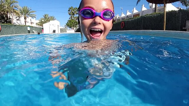 Cheerful pretty little 9s adorable girl wear swimsuit goggles looking at camera enjoy summer vacations swimming in pool. Active lifestyle and sport activity, summertime pleasure concept. Slow motion