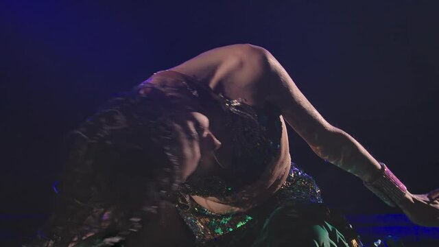 Wet oriental woman sitting on the surface of the water and dancing a belly dance. An exotic dancer with long black hair in a traditional costume performs in a dark studio. Slow motion. Close up.