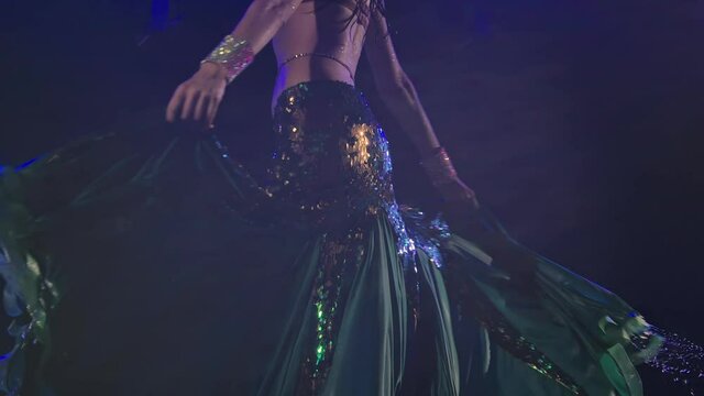 Bottom view of young oriental dancer dancing and swinging a long green skirt. Woman dances on the surface of the water creating splashes in a dark studio. Close up slender legs . Slow motion.