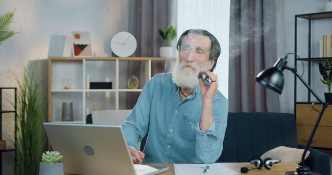 Handsome relaxed positive bearded old man with wrinkled face sitting in front of camera and smoking a cigar with pleasure on contemporary room interier background