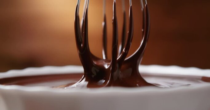 Close-up mixing melted liquid chocolate with steel whisk in kitchen. Hot chocolate mix swirl and drip in white bowl on wooden background. Cooking handmade chocolate dessert. Confectionery concept