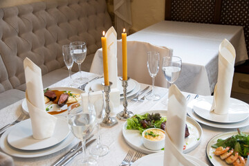 Romantic candlelight dinner in a beautiful restaurant. Natural wax candles on the table. Lighted candles. Side view on the served banquet restaurant table with candles, wine glasses, napkins, plates.