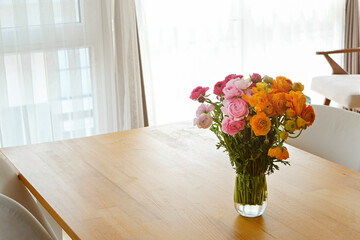 Close up shot of wooden dining table with beautiful ranunculuses in glass vase. A bouquet of mixed persian buttercup flowers in soft natural light from a window. Copy space, close up, background.