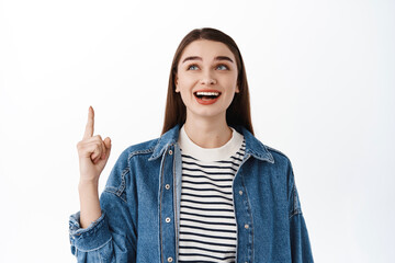 Smiling caucasian female model pointing, looking up with happy pleased face, checking out promotional offer, logo or banner on top copyspace, standing over white background