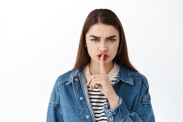 Angry young woman shushing, frowning and make shhh hush gesture with finger on lips, taboo sign,...