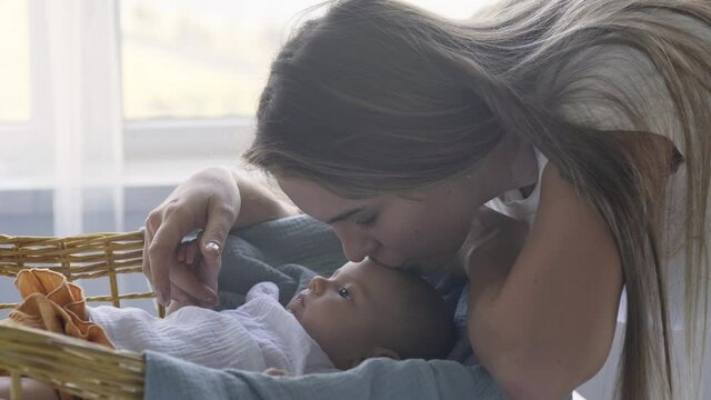 Long haired mother plays with baby and kisses kid forehead sitting at wooden wicker cradle against large window with bright light close-up