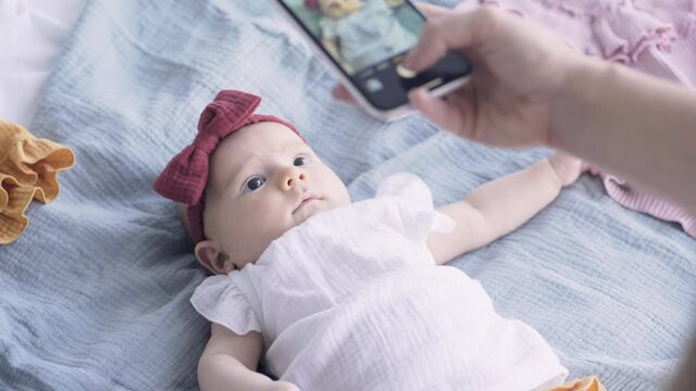 Mother hand makes photos with black smartphone of funny baby daughter with bow lying on large bed under bright light at home in morning close view