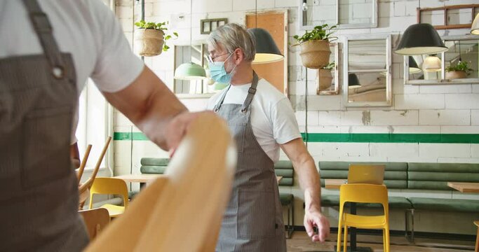 Portrait Of Caucasian Old Senior Male Manager In Medical Mask Walking In Restaurant Telling Mixed-race Young Workers What To Do. Cleaning Cafeteria At Workplace In Quarantine. Coronavirus, Business