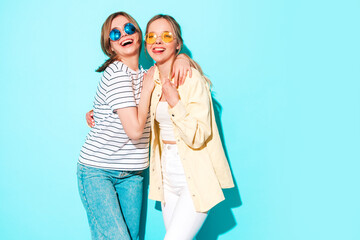 Two young beautiful blond smiling hipster female in trendy summer clothes. Sexy carefree women posing near blue wall in studio. Trendy and positive models having fun in sunglasses