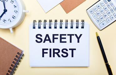 On a light background, a white alarm clock, a calculator, a pen and a notebook with the text SAFETY FIRST. Flat lay