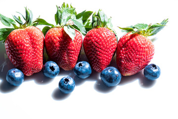 Four very red strawberries with their green leaves and several blueberries on a white background.