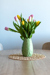 Vertical photo of a beautiful composition of pink and yellow tulips inside a green vase on a wooden table. White background.