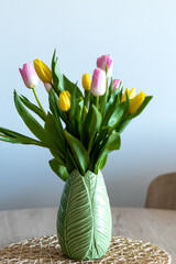 Vertical photo of a beautiful composition of pink and yellow tulips inside a green vase on a wooden table. White background and selective focus.