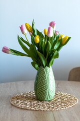 Vertical photo of a nice composition of pink and yellow tulips inside a green vase on a wooden table inside my house. White background and selective focus.