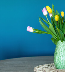 Vertical photo of pink and yellow tulips inside a green vase inside my house. Composition of half vase. Oil blue background.
