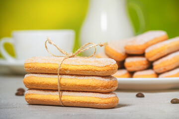 Italian savoiardi biscuits or ladyfingers cookies on a plate and in a stack, a white cup and a jug of milk in the background. Selective focus
