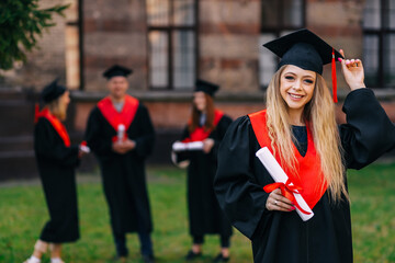 Portrait of a confident female student with a diploma in hands on the background of the university. The girl is dressed in mortar boards and a robe. Education, graduation and people concept.