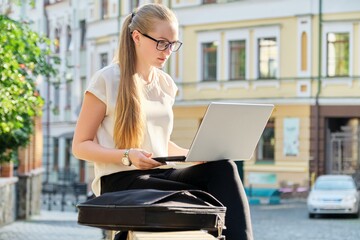 Beautiful young woman sitting outdoors using laptop, city street background