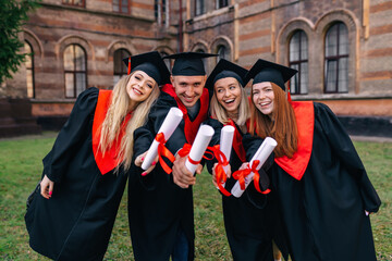 Successful graduates in academic gowns hold their diplomas, look at the camera and smile for an...