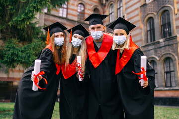 A graduate wearing medical masks on his face. Happy students with diplomas in an academic gown and graduation cap, a group with a certificate of education, are standing near the university.