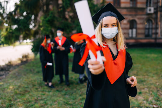 High School Graduation. Beautiful Female Graduate In A Medical Protective Mask On Her Face And A Hat On Her Head, Outdoors, Shows A Diploma With A Red Ribbon. Distance Learning, Coronavirus.