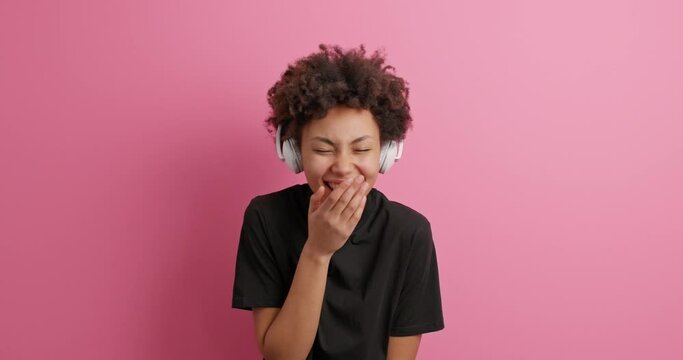 Positive upbeat dark skinned beautiful woman cannot stop laughing being amused by someone hears something funny wears wireless headphones on ears dressed casually isolated over pink background
